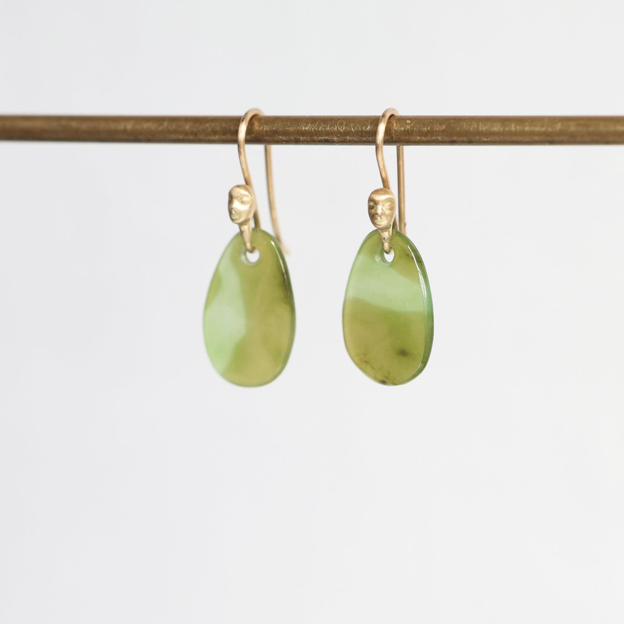 A pair of gold-tone drop earrings with polished light green teardrop-shaped stone pendants, hanging from a horizontal gold rod against a plain white background.
