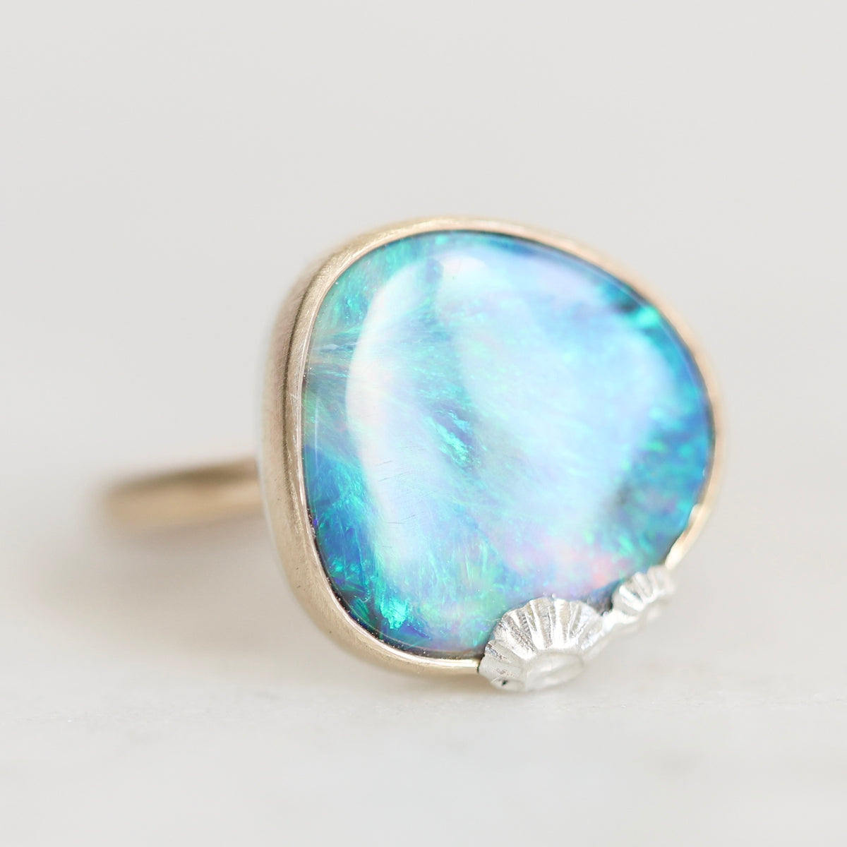 A silver ring featuring a large, irregularly shaped cabochon opal gemstone with iridescent blue, green, and white play-of-color, accented by a small shell-shaped detail on the lower right edge of the stone.