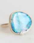 A silver ring featuring a large, irregularly shaped cabochon opal gemstone with iridescent blue, green, and white play-of-color, accented by a small shell-shaped detail on the lower right edge of the stone.