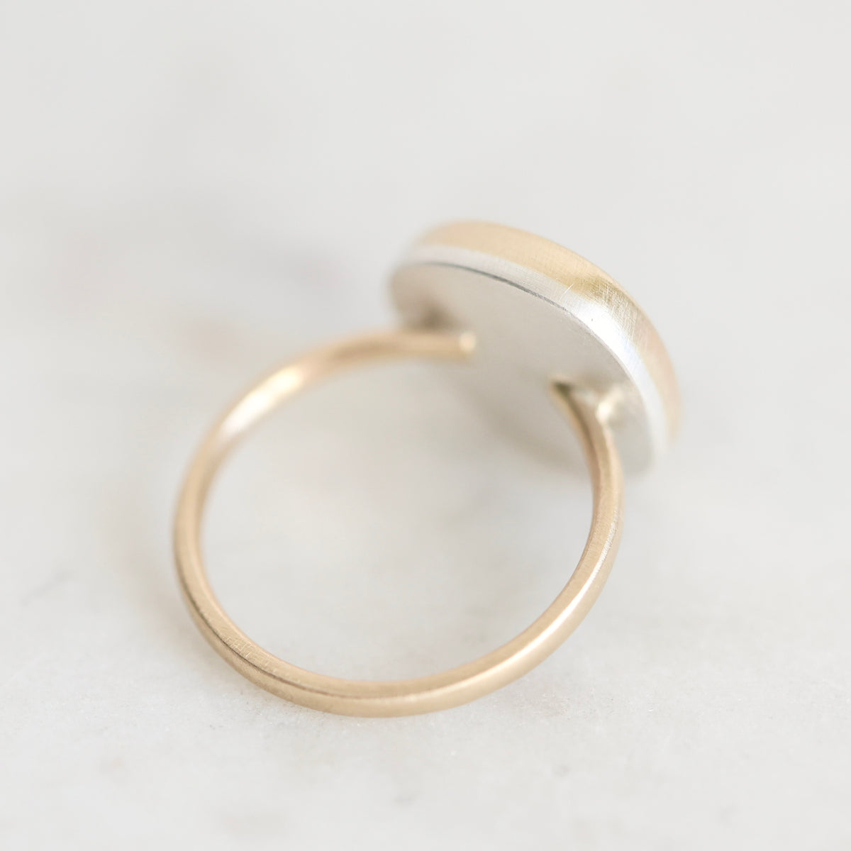 A minimalist gold ring with a flat, circular silver-toned top, photographed on a light gray marble surface.