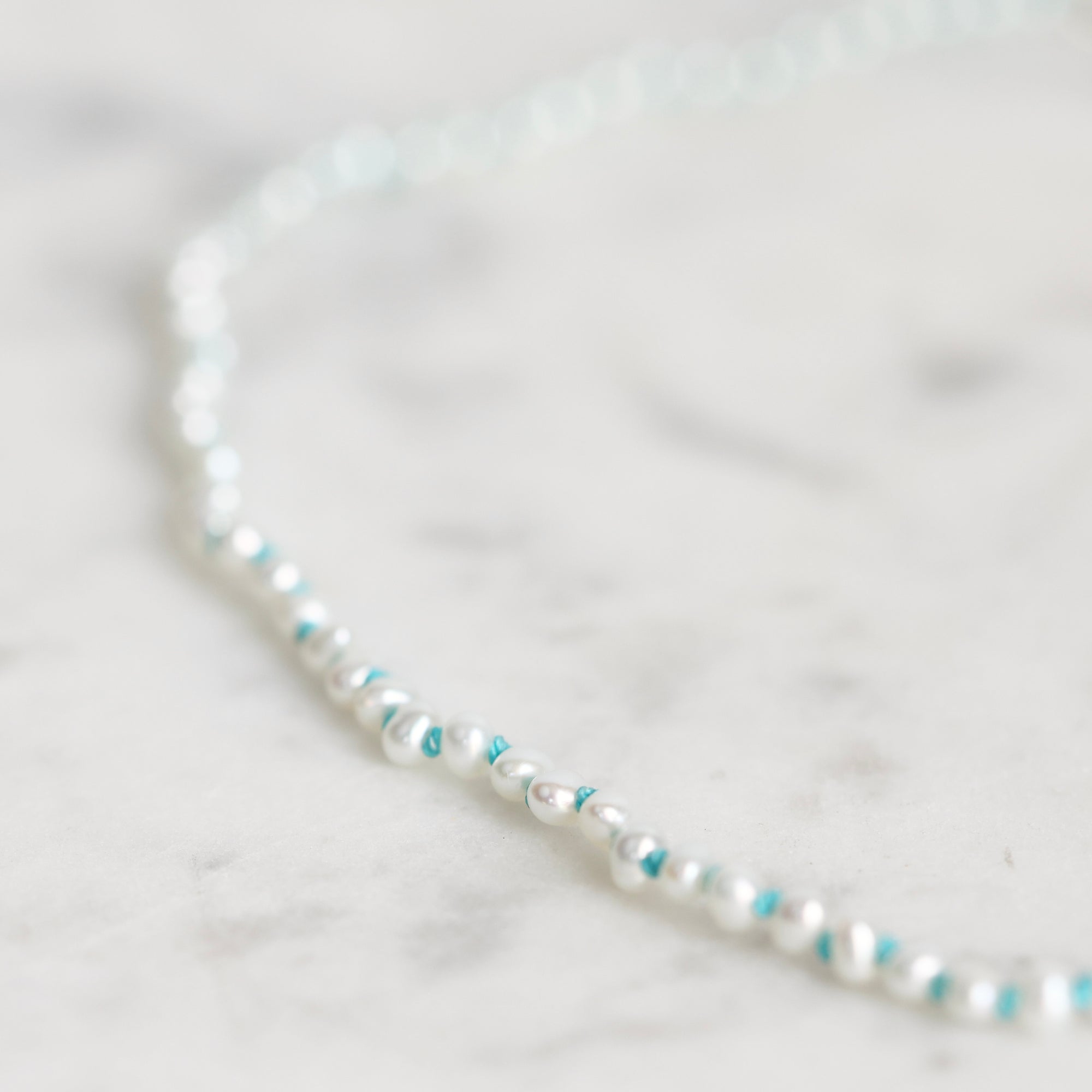 A delicate beaded necklace with white pearl-like beads and small turquoise blue accent beads, photographed on a light marble surface.