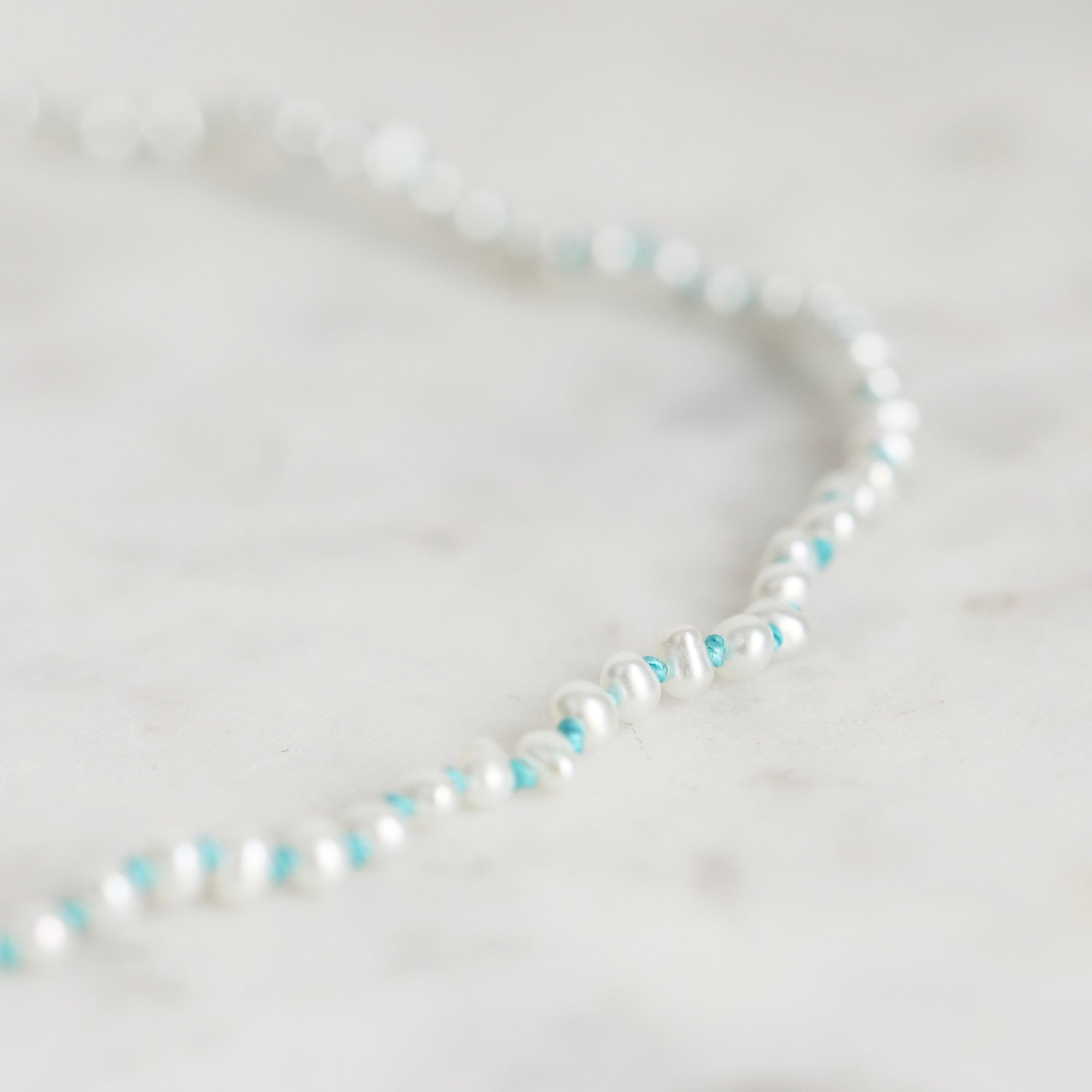 A close-up of a delicate beaded strand necklace with white pearl-like beads and small turquoise blue accents, resting on a light marble surface.