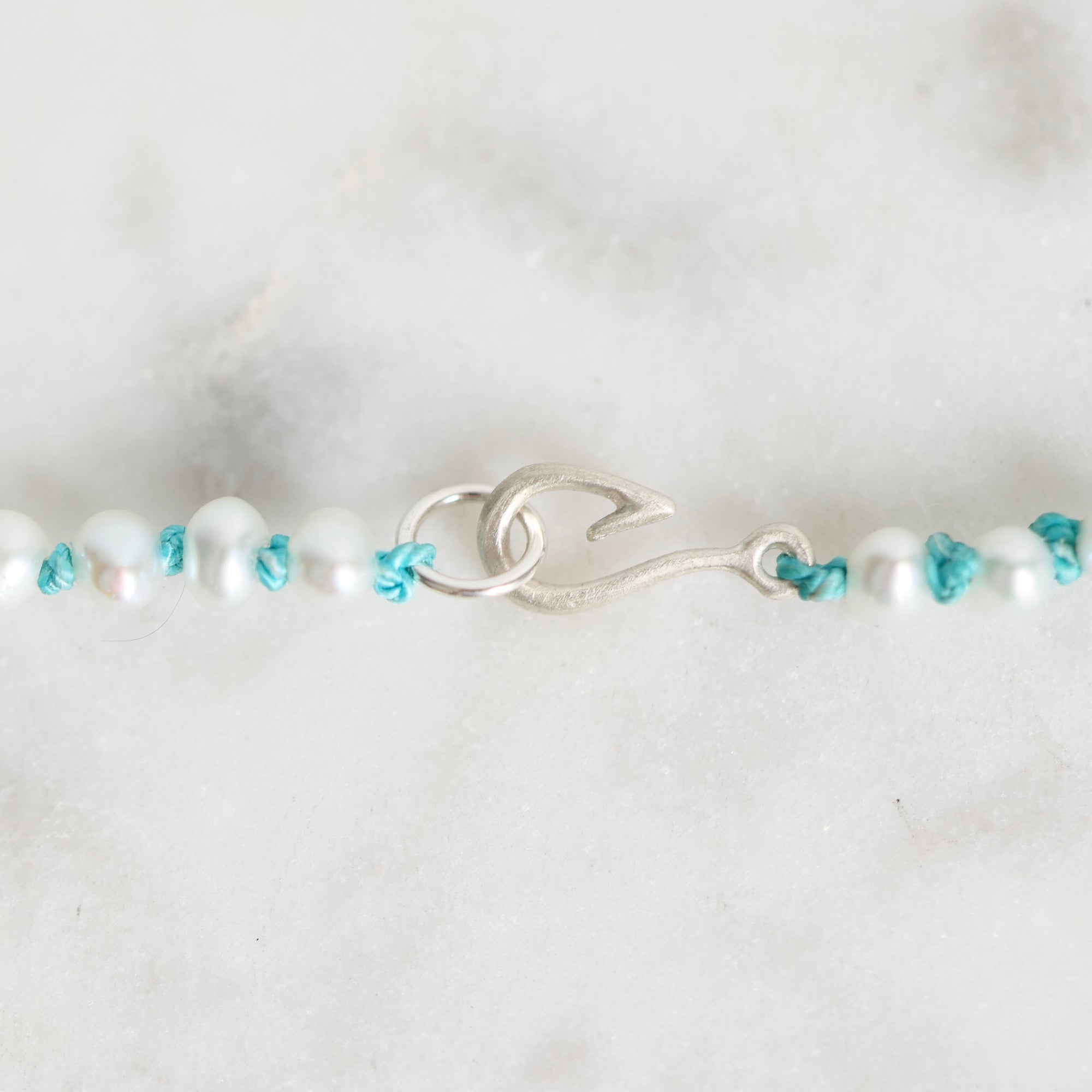 A close-up of a beaded bracelet featuring white pearl-like beads, turquoise blue knotted accents, and a textured silver clasp, displayed on a light marble surface.