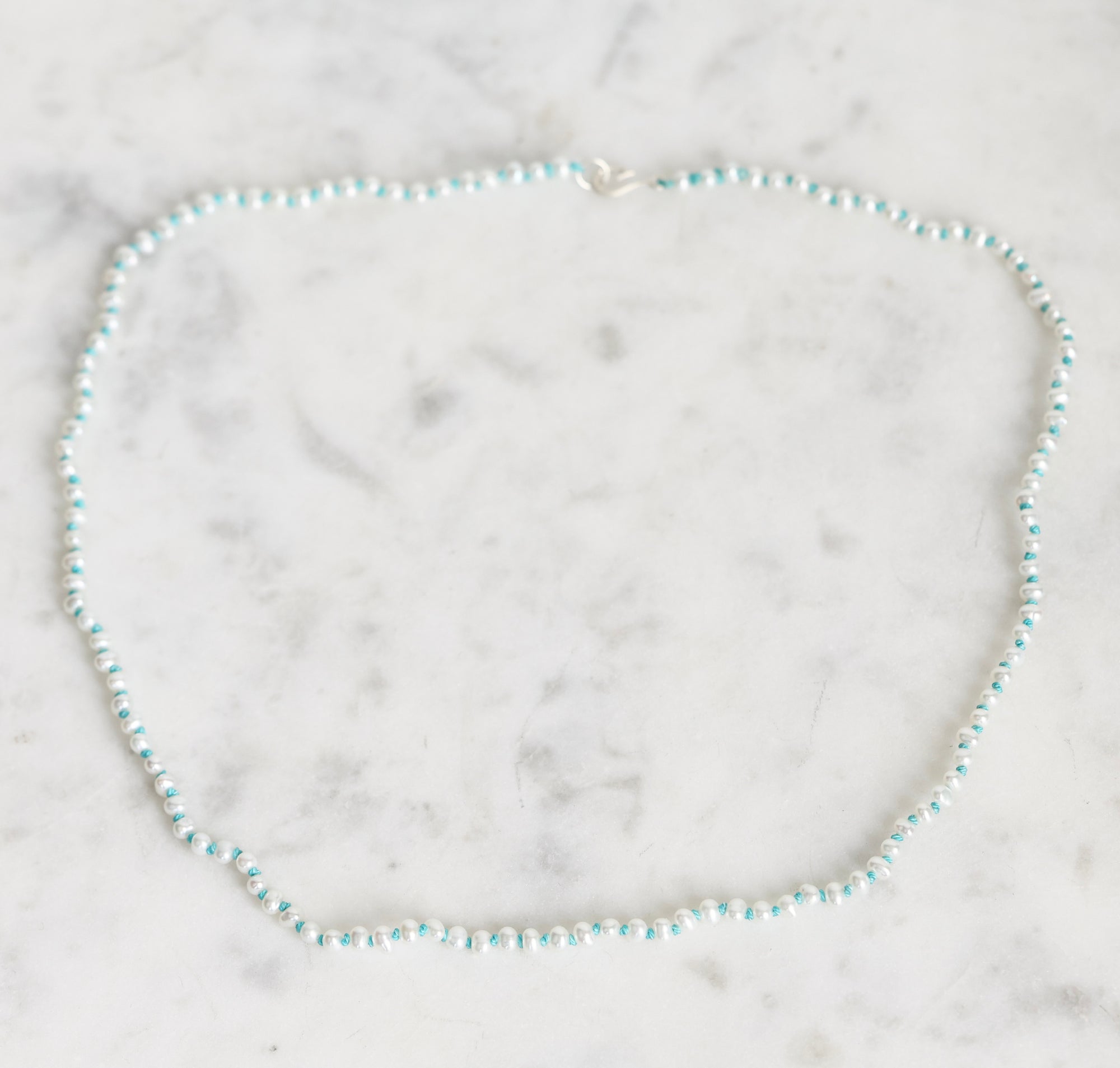 A delicate beaded necklace with alternating light blue and white beads, arranged in a circle on a white marble surface.