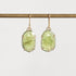 A pair of oval green gemstone drop earrings in gold-toned prong settings hanging from gold-toned hooks, suspended from a horizontal gold rod against a light gray background.