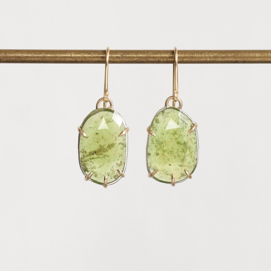 A pair of oval green gemstone drop earrings in gold-toned prong settings hanging from gold-toned hooks, suspended from a horizontal gold rod against a light gray background.