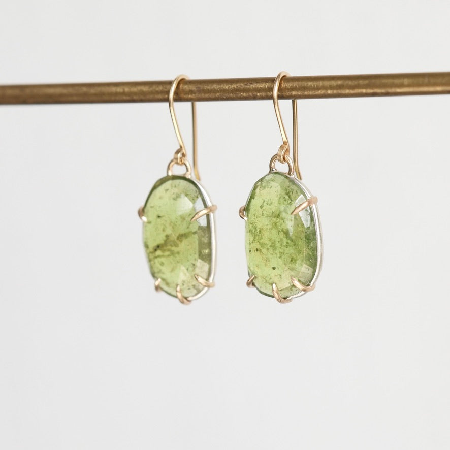 A pair of oval green stone drop earrings in gold-toned settings, hanging from a gold-toned horizontal bar against a plain light gray background.