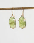 A pair of oval green stone drop earrings in gold-toned settings, hanging from a gold-toned horizontal bar against a plain light gray background.
