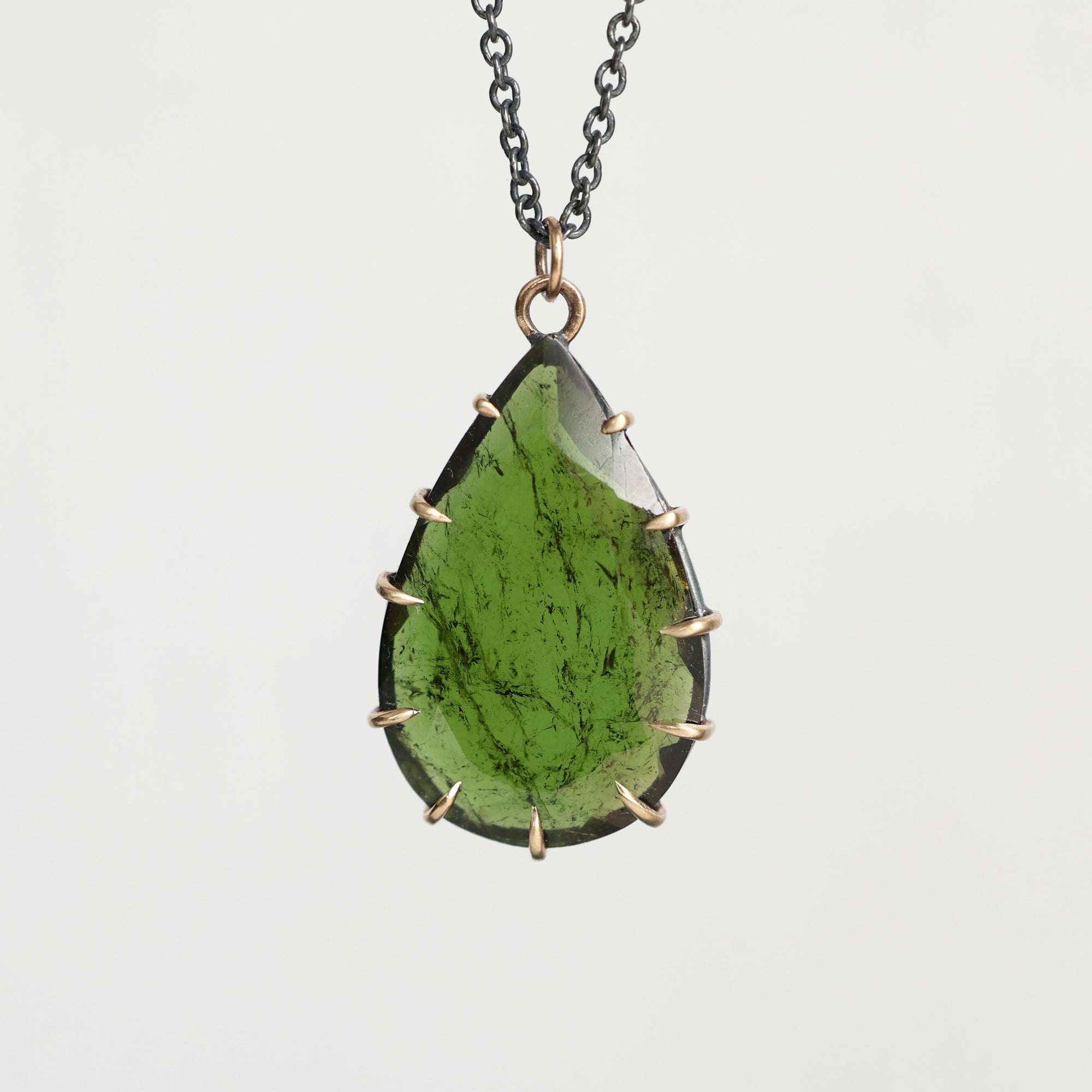 A teardrop-shaped green gemstone pendant necklace with a dark metal chain and a bronze-toned bezel setting, shown against a light gray background.
