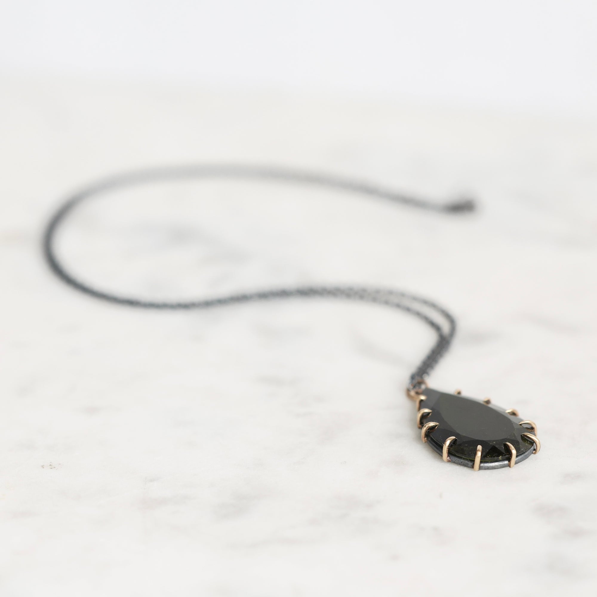 A black teardrop pendant necklace with a gold-toned spiked border, resting on a white marble surface.