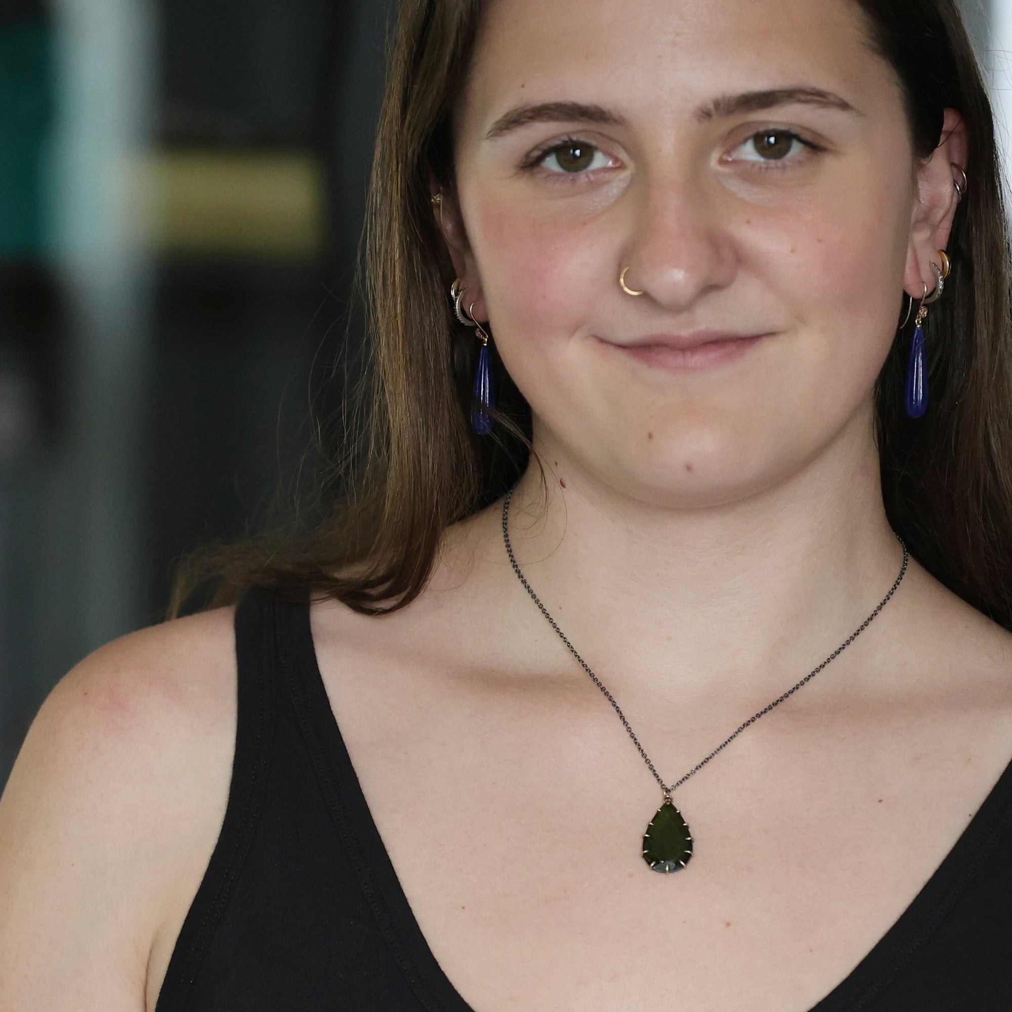 A woman wearing a dark green teardrop pendant necklace on a black chain, paired with matching dark blue teardrop earrings, shown in a close-up portrait.