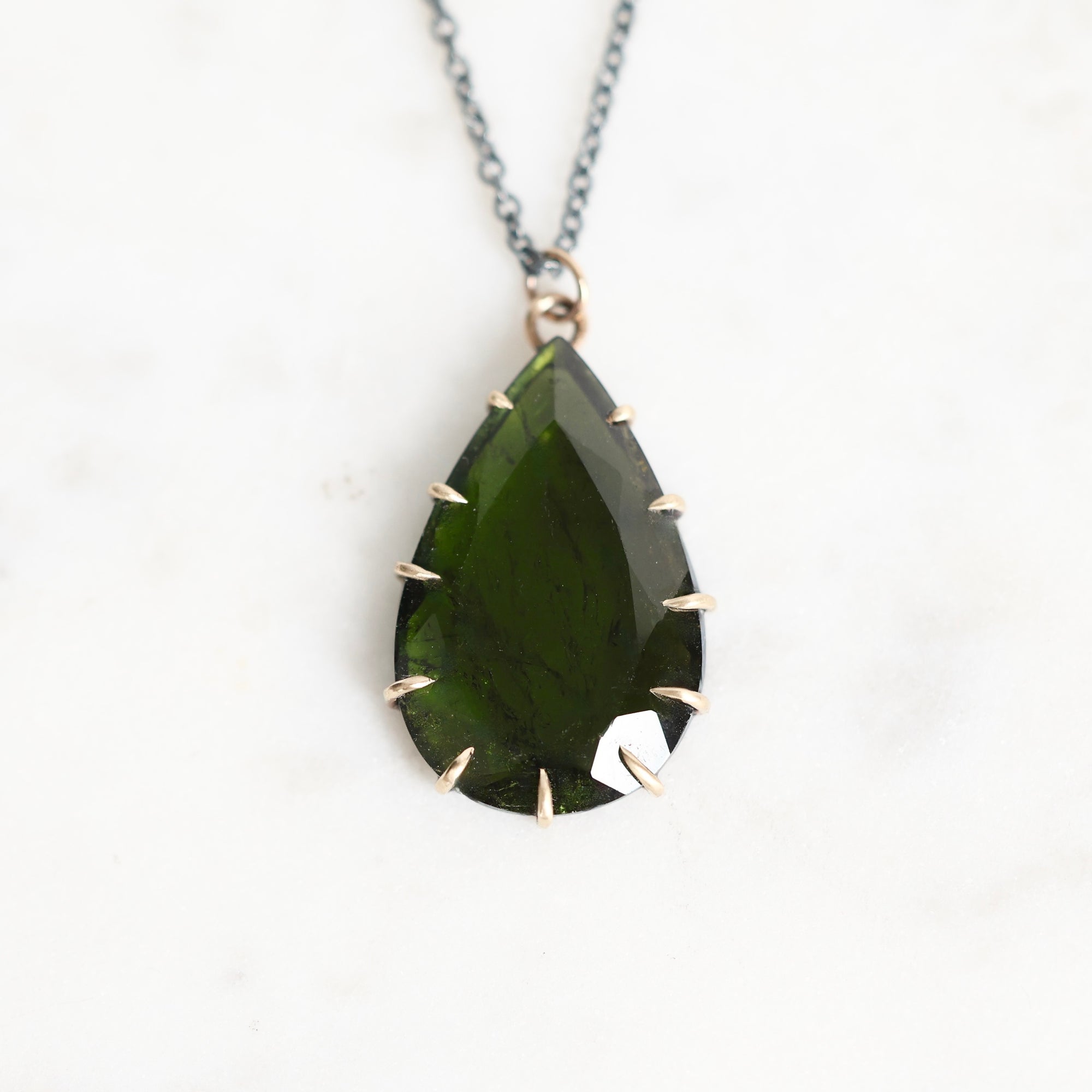 A teardrop-shaped dark green gemstone pendant with gold-toned prong settings on a silver-toned chain, displayed on a light marble background.