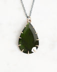 A teardrop-shaped dark green gemstone pendant with gold-toned prong settings on a silver-toned chain, displayed on a light marble background.