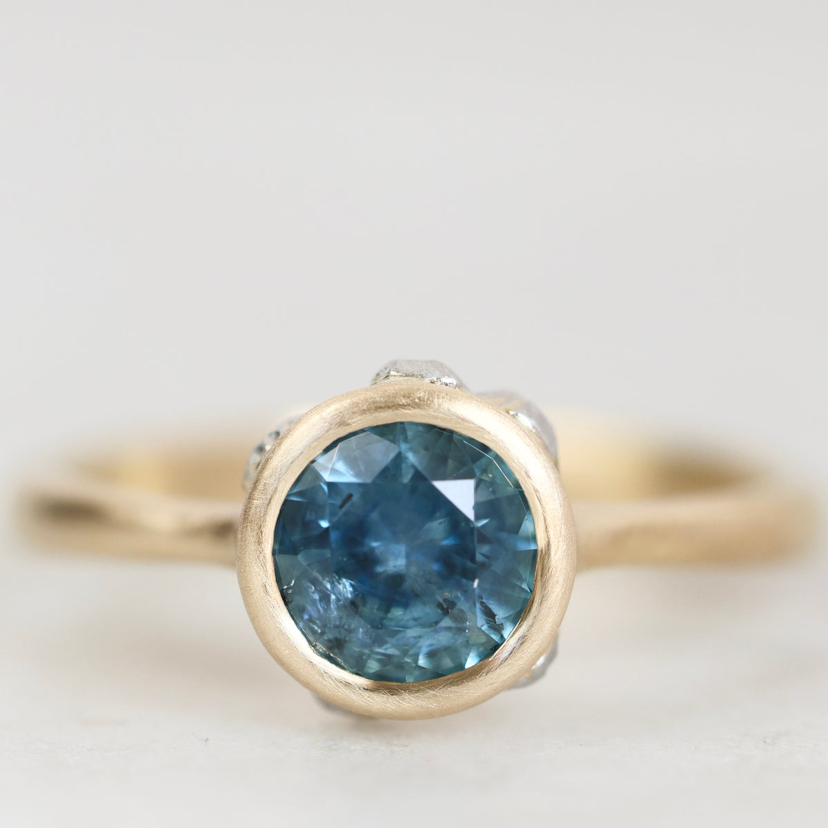A close-up shot of a gold ring featuring a round-cut blue gemstone, set against a soft, light gray background.