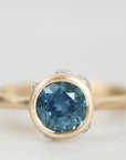 A close-up shot of a gold ring featuring a round-cut blue gemstone, set against a soft, light gray background.