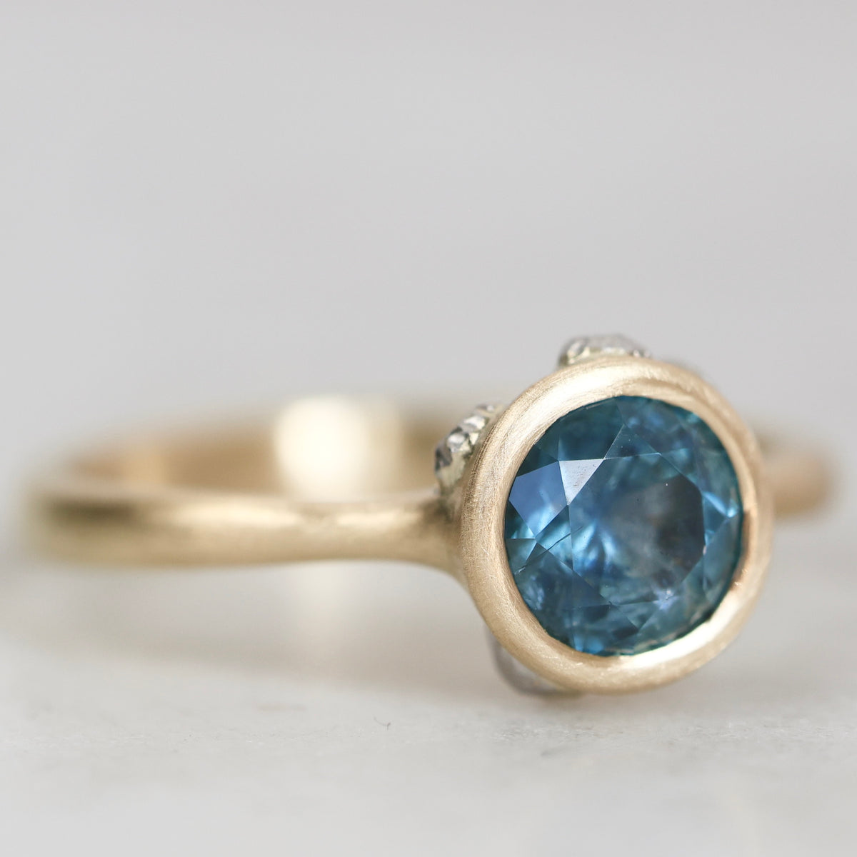 A gold ring featuring a round-cut blue gemstone in a bezel setting, with small accent stones on either side of the main stone, photographed on a light gray background.