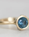 A gold ring featuring a round-cut blue gemstone in a bezel setting, with small accent stones on either side of the main stone, photographed on a light gray background.