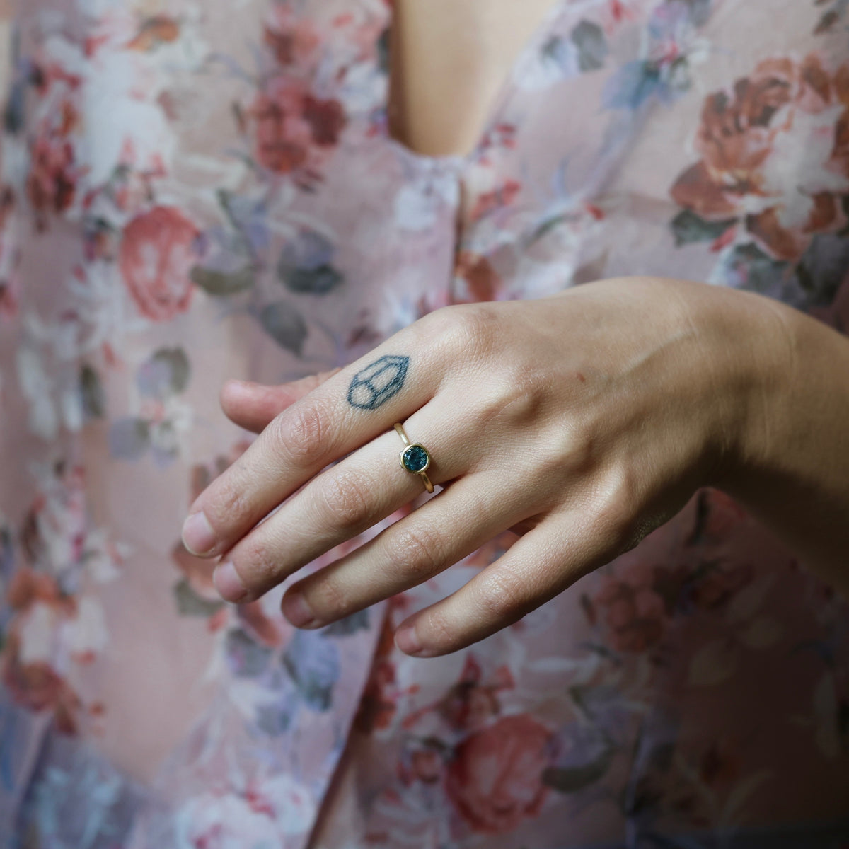 Woman's hand with tattoo posed wearing bezel set blue stone ring against a floral blouse. 
