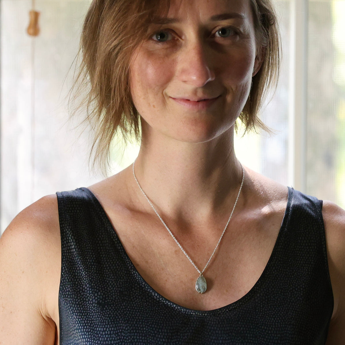 A woman with short light brown hair wearing a silver chain necklace with a teardrop-shaped gray stone pendant, paired with a black textured tank top.