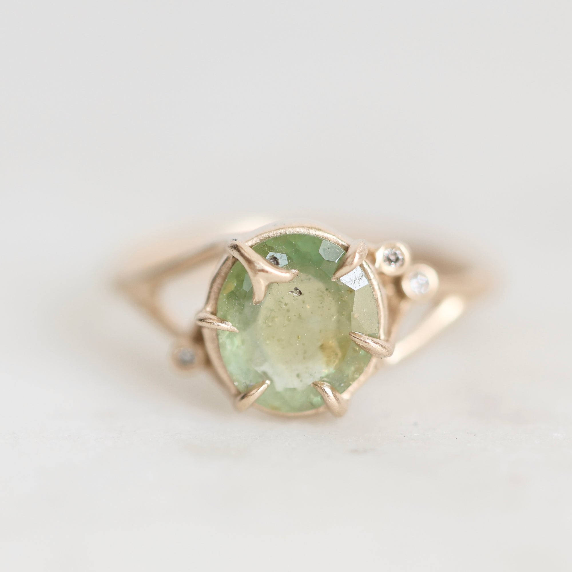 A yellow gold ring featuring a round-cut light green gemstone in a prong setting, accented with small clear stones on the band, shown on a light gray background.