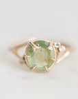 A yellow gold ring featuring a round-cut light green gemstone in a prong setting, accented with small clear stones on the band, shown on a light gray background.