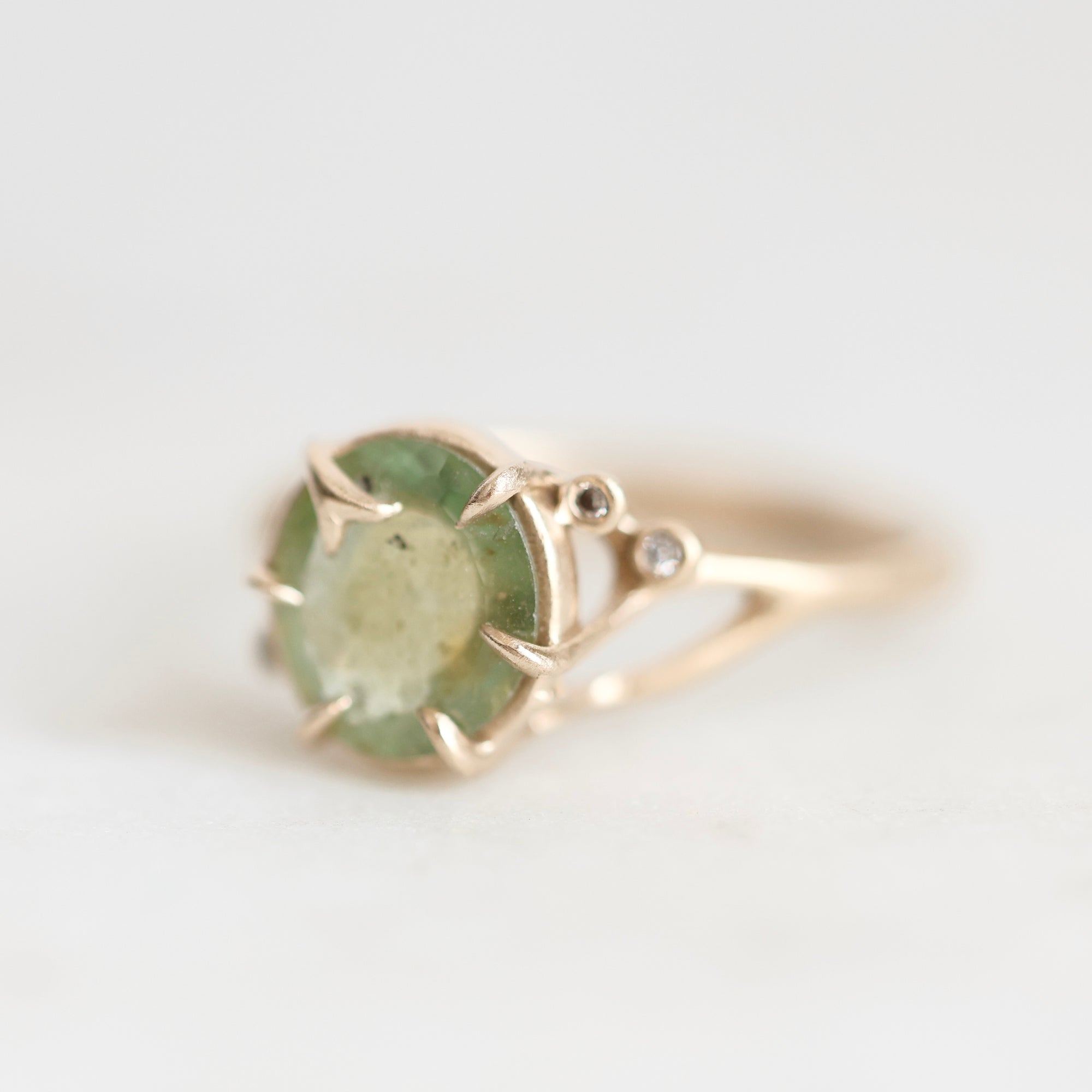 A gold ring featuring a round, faceted light green gemstone in a prong setting, with a small clear accent stone on the band, photographed against a soft white background.