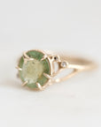 A gold ring featuring a round, faceted light green gemstone in a prong setting, with a small clear accent stone on the band, photographed against a soft white background.