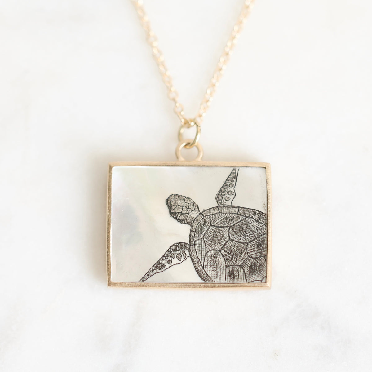 A rectangular pendant necklace featuring a mother-of-pearl inlay with a black engraved sea turtle design, suspended from a delicate gold chain on a light marble background.