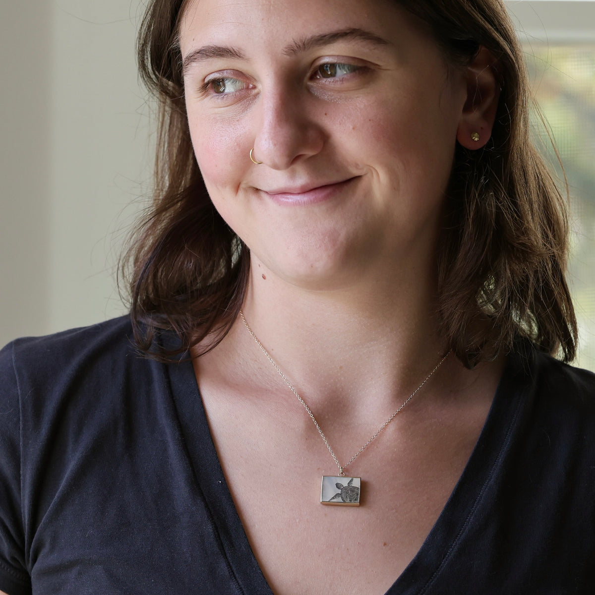 A woman wearing a silver-toned rectangular pendant necklace with a floral engraving, paired with a dark navy blue v-neck t-shirt.