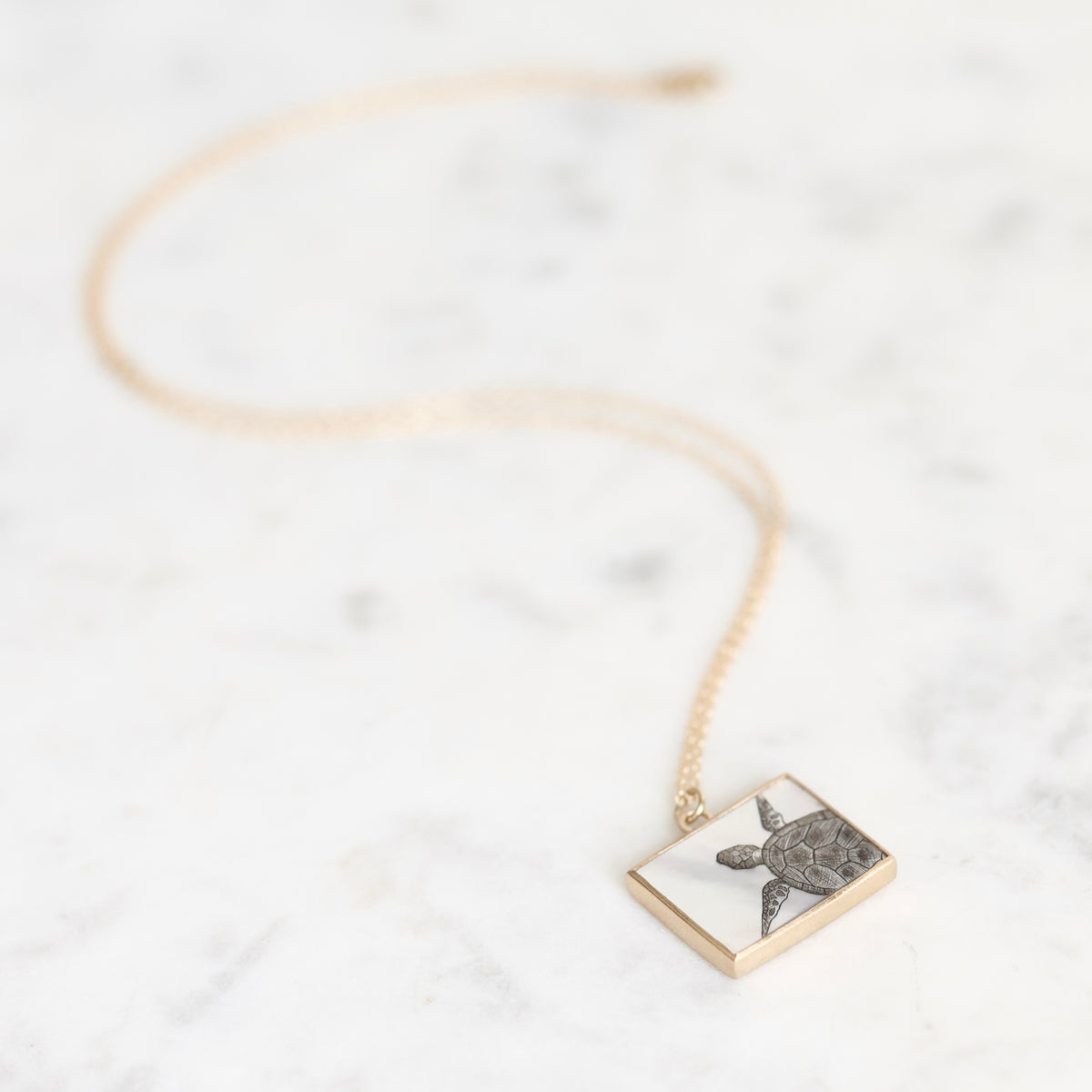 A gold chain necklace with a square pendant featuring a silver-toned sea turtle illustration, displayed on a white marble surface.