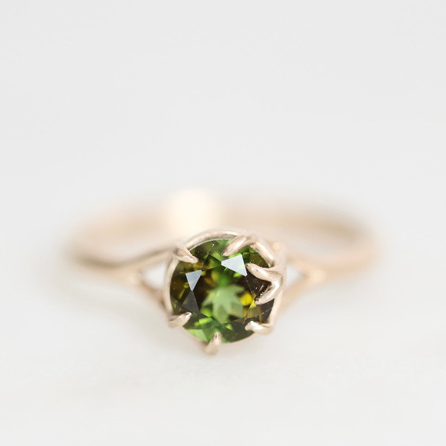 A gold ring featuring a round green gemstone in a prong setting, photographed against a soft, light gray background.
