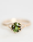 A gold ring featuring a round green gemstone in a prong setting, photographed against a soft, light gray background.
