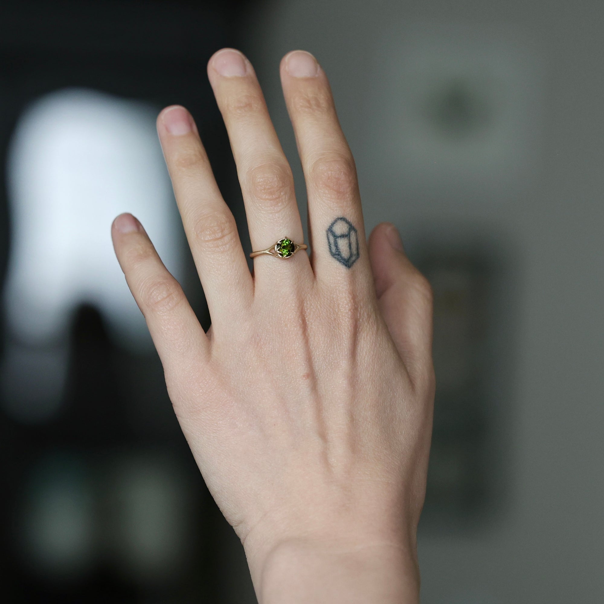 A close-up of a person's hand wearing a delicate gold ring with a small green gemstone, set against a softly blurred dark background.