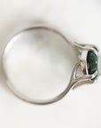 A silver ring with a marquise-cut green gemstone, shown in a close-up side view on a soft white background.
