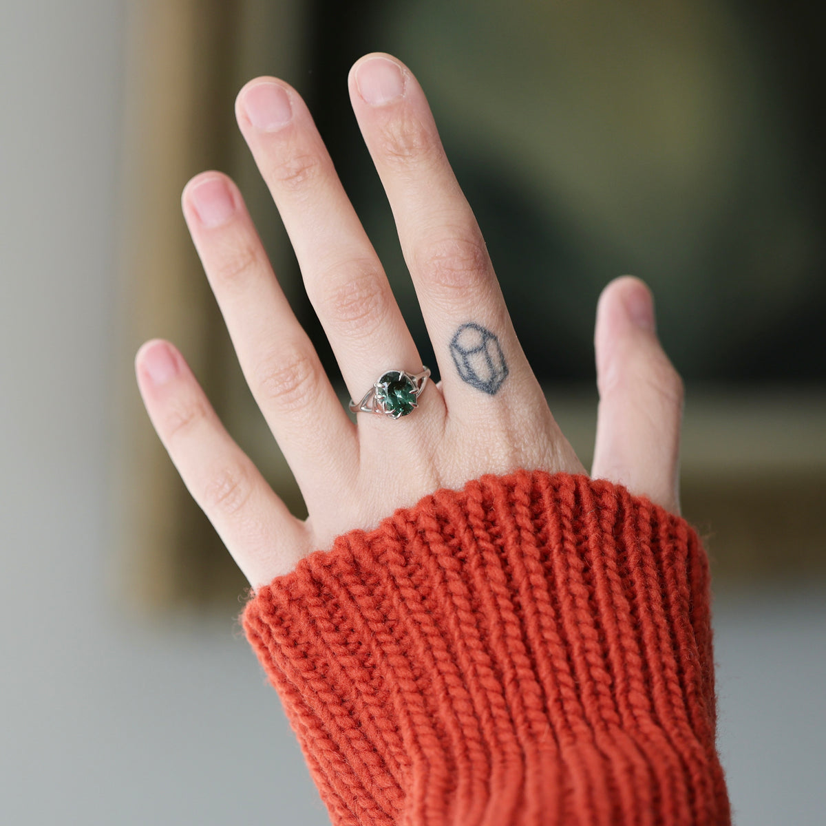 A close-up of a person's hand wearing a silver ring with a green faceted gemstone, styled with a rust-orange ribbed knit sweater sleeve. The background is a soft, blurred indoor setting.