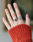 A close-up of a person's hand wearing a silver ring with a green faceted gemstone, styled with a rust-orange ribbed knit sweater sleeve. The background is a soft, blurred indoor setting.
