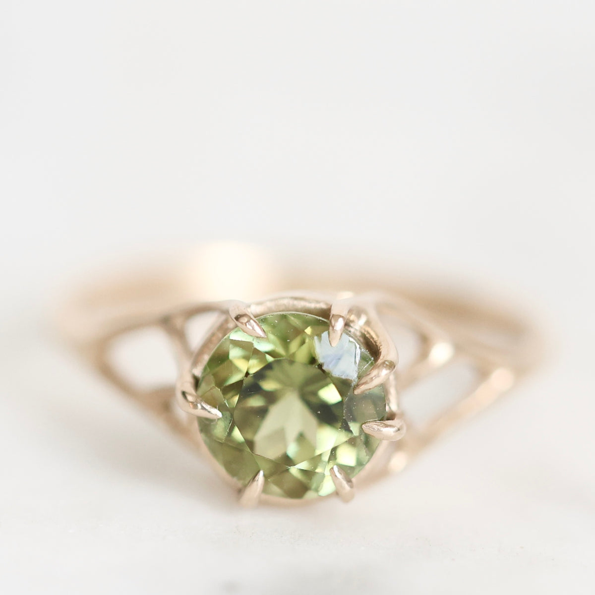 A close-up shot of a ring featuring a round faceted green gemstone in a prong setting on a gold-toned band, set against a soft, out-of-focus light background.