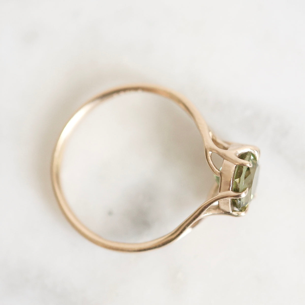 A close-up shot of a gold ring with a green rectangular gemstone, resting on a light marble surface.