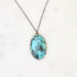 An oval pendant necklace featuring a turquoise-blue stone with dark matrix inlay, set in a rose gold-toned prong setting on a silver-toned chain, displayed on a white marble background.