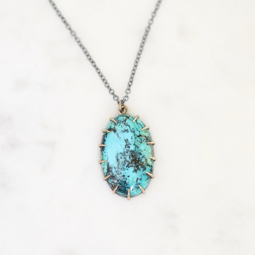 An oval pendant necklace featuring a turquoise-blue stone with dark matrix inlay, set in a rose gold-toned prong setting on a silver-toned chain, displayed on a white marble background.