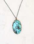 An oval pendant necklace featuring a turquoise-blue stone with dark matrix inlay, set in a rose gold-toned prong setting on a silver-toned chain, displayed on a white marble background.