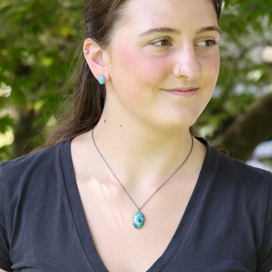 A woman wearing a silver chain necklace with a turquoise-colored oval pendant, photographed outdoors with a blurred green background.