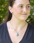 A woman wearing a silver chain necklace with a turquoise-colored oval pendant, photographed outdoors with a blurred green background.