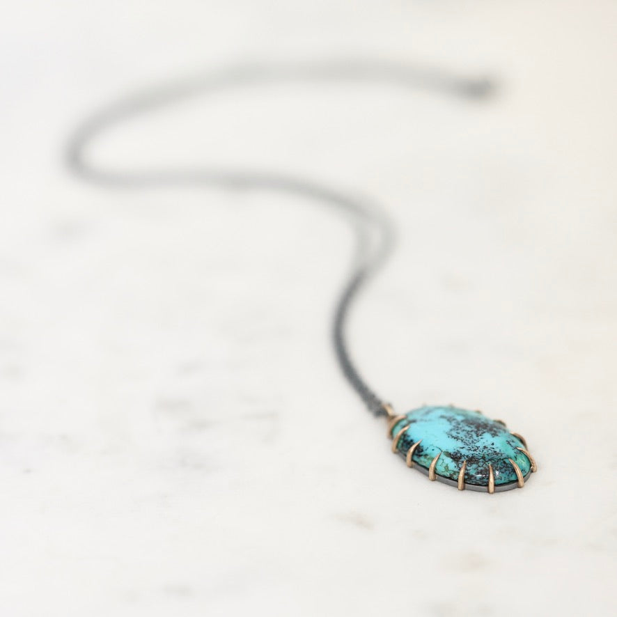 A close-up shot of a turquoise pendant necklace with a textured, hammered finish, set in a gold-toned bezel and suspended from a dark cord, resting on a light marble surface.