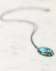 A close-up shot of a turquoise pendant necklace with a textured, hammered finish, set in a gold-toned bezel and suspended from a dark cord, resting on a light marble surface.