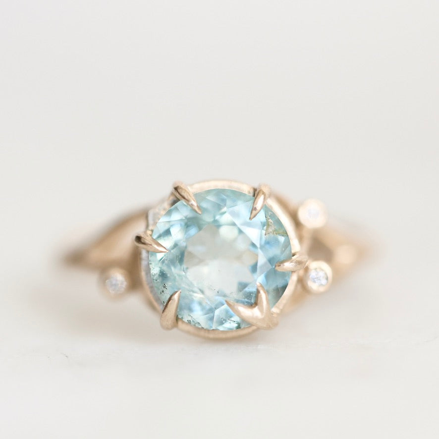 A gold ring featuring a large round-cut light blue gemstone in a prong setting, with small accent stones on the band, photographed against a soft white background.
