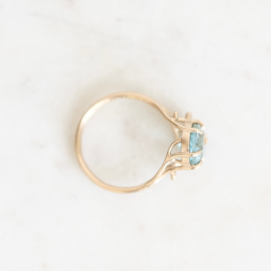 A delicate yellow gold ring featuring a small, round-cut light blue gemstone in a prong setting, photographed from above on a light marble surface.