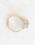 A delicate yellow gold ring featuring a small, round-cut light blue gemstone in a prong setting, photographed from above on a light marble surface.