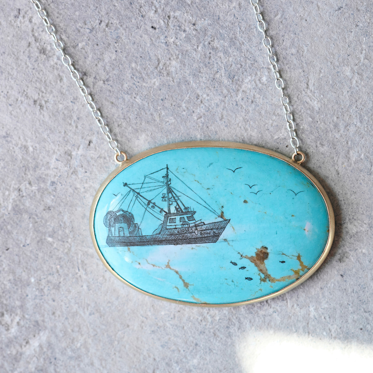 An oval turquoise pendant necklace featuring a hand-painted black and white ship illustration, set in a gold-tone frame on a silver chain, displayed on a textured gray stone surface.