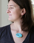 Woman wearing large turquoise pendant necklace looking out the window against a blurred indoor background.